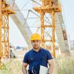 Engineer in safety gear holding a clipboard at a construction site with arches in the background.